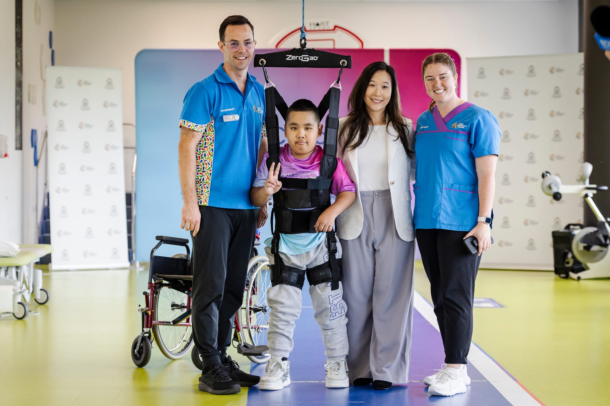 A child using a body‑support walking harness stands on a therapy floor with his mother and two healthcare staff members; a wheelchair is positioned behind.