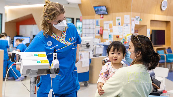A nurse discusses patient care with a mother, who is holding her toddler daughter who is facing the camera