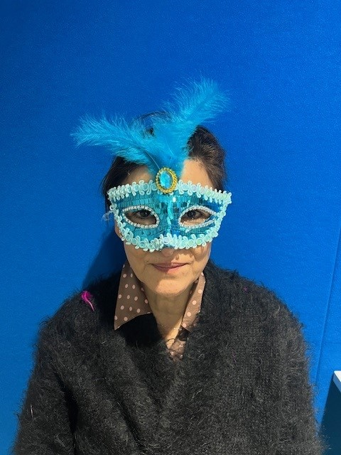 A mystery doctor concealing their identity by holding a mask over their face. The doctor is wearing a navy-blue scrub top and maroon scrub hat.